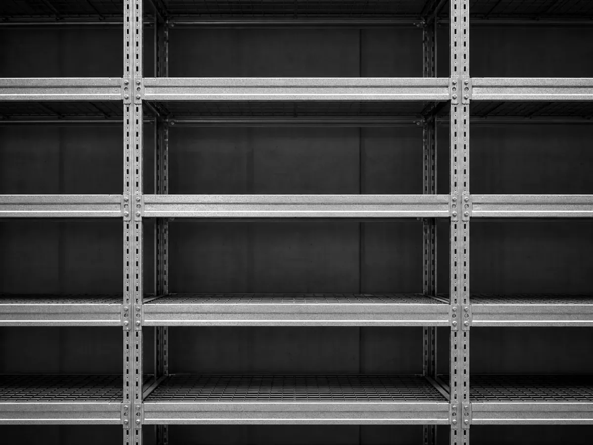 A frontal close-up of a galvanized steel pallet racking system in a warehouse — vertical uprights and horizontal beams forming a uniform grid, the steel surface showing fine workshop texture.