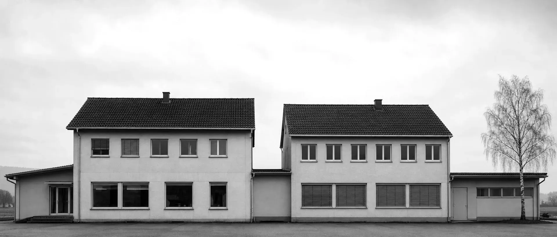 A frontal typology shot of two adjacent plain Swiss commercial buildings in flat Mittelland daylight: straight rendered facades, evenly spaced windows, empty grey sky above.