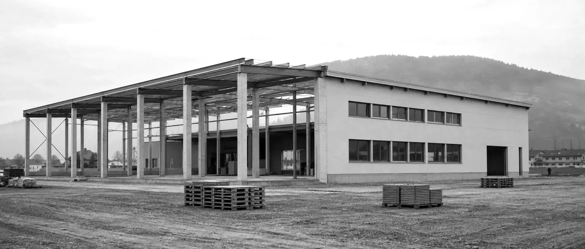 Eine ruhige Weitwinkelaufnahme einer schlichten Schweizer Industriehalle im Spätstadium des Baus: sichtbare Betonpfeiler, teilverputzte Fassade, flacher Bauplatz im Vordergrund, weicher bewölkter Himmel.