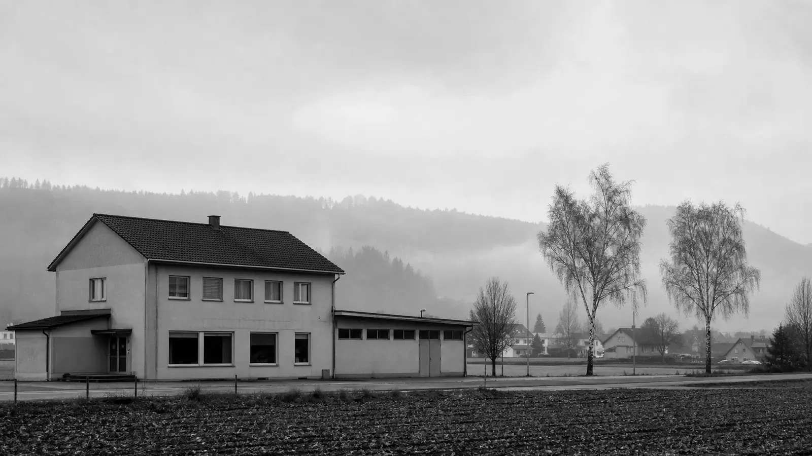 A plain two-storey commercial building in a small Seeland village in early morning, light mist over flat farmland, gently wooded Jura hills in the distance — rural canton of Bern.